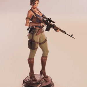 May include: A detailed figurine of a woman holding a rifle, dressed in a blue top, green pants, and brown boots. The figure stands on a base with a red and black design, and a weapon. The figurine has a serious expression.