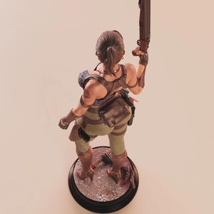 May include: A detailed figurine of a female character in combat gear, holding a shotgun. The figure is posed on a circular base with a detailed, textured surface. The character wears a brown leather harness, green pants, and brown boots.
