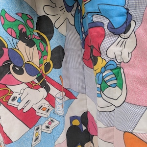 K&ouml;nnte beinhalten: Ein farbenfroher Stoff mit einem sich wiederholenden Muster von Mickey und Minnie Mouse in verschiedenen Posen. Minnie tr&auml;gt Sonnenbrille und Kopfh&ouml;rer. Das Design umfasst Kassetten, ein Getr&auml;nk und geometrische Formen in Rot, Blau und Rosa.