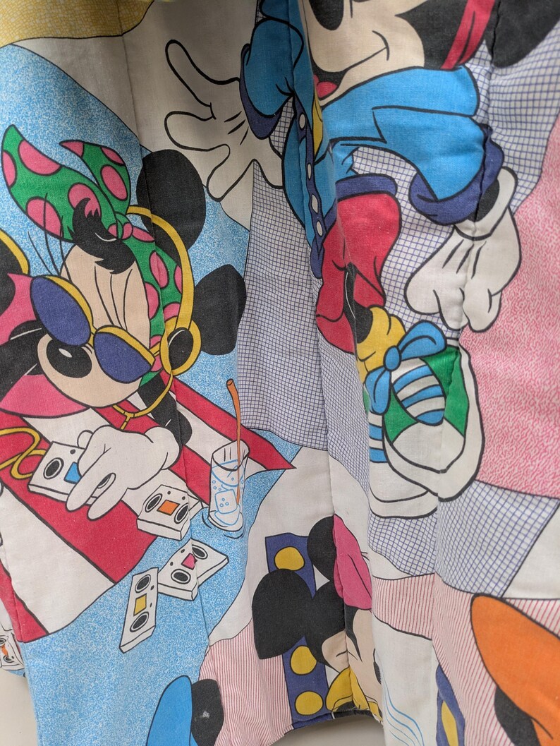 K&ouml;nnte beinhalten: Ein farbenfroher Stoff mit einem sich wiederholenden Muster von Mickey und Minnie Mouse in verschiedenen Posen. Minnie tr&auml;gt Sonnenbrille und Kopfh&ouml;rer. Das Design umfasst Kassetten, ein Getr&auml;nk und geometrische Formen in Rot, Blau und Rosa.