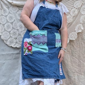 May include: A denim apron with patchwork pockets. The pockets feature floral and geometric patterns in shades of pink, purple, and teal. The apron is worn over a white top.