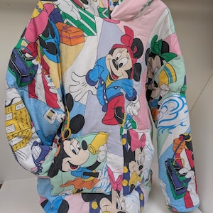K&ouml;nnte beinhalten: Ein Patchwork-Hoodie mit Disney-Charakterdrucken, darunter Mickey und Minnie Mouse. Der Hoodie hat eine Kapuze, lange &Auml;rmel und eine Vordertasche. Das Design beinhaltet eine Vielzahl von Farben und Mustern.