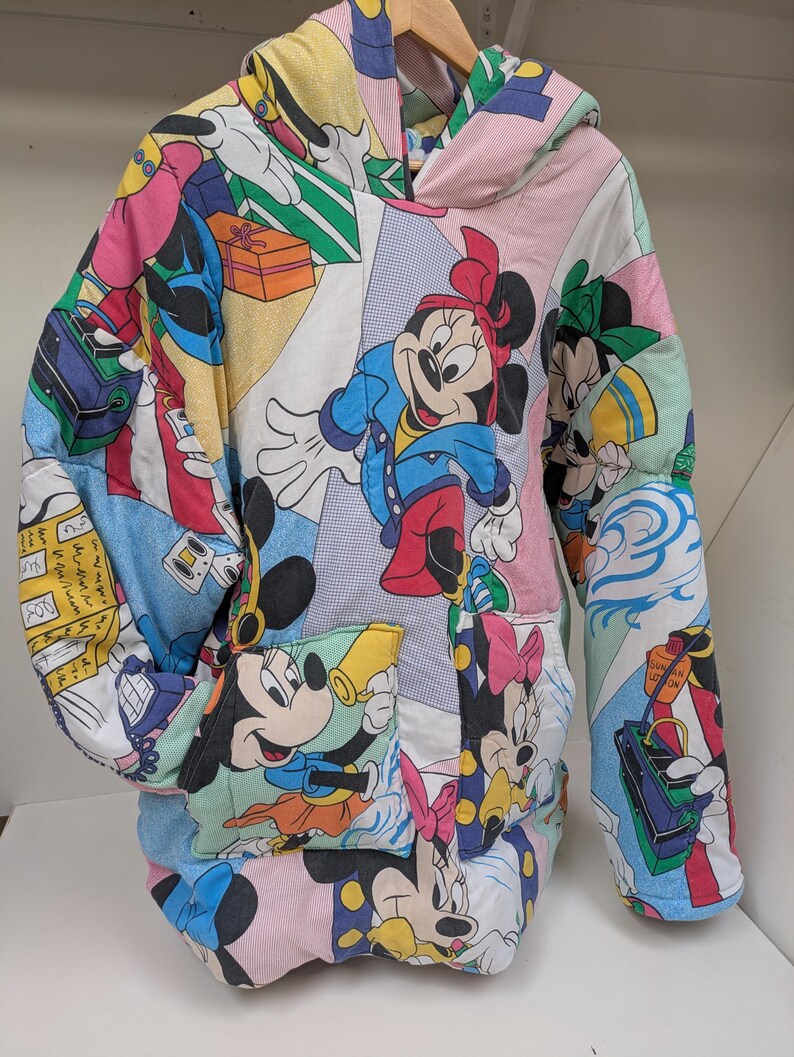 K&ouml;nnte beinhalten: Ein Patchwork-Hoodie mit Disney-Charakterdrucken, darunter Mickey und Minnie Mouse. Der Hoodie hat eine Kapuze, lange &Auml;rmel und eine Vordertasche. Das Design beinhaltet eine Vielzahl von Farben und Mustern.