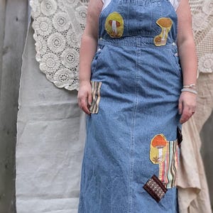 May include: A denim overall dress with mushroom patches. The dress is a light blue wash with a long skirt and adjustable straps. Patches of mushrooms and colorful fabric are sewn on the front. The dress is worn over a white top.