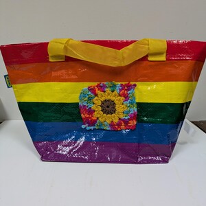 May include: A rainbow-striped tote bag with yellow handles and a crocheted sunflower applique. The bag features a vibrant spectrum of colors, including red, orange, yellow, green, blue, and purple. The sunflower applique adds a handcrafted touch.
