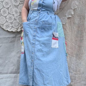 May include: Light blue denim overall dress with floral and striped patchwork details. The dress features a button-down front, large front pockets, and adjustable straps. The dress is knee-length and has a relaxed fit.