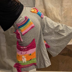 May include: Grey sweatpants featuring vibrant, knitted patches. The patches showcase rainbow stripes, a lamb, and other playful designs. These patches are sewn onto the trousers, giving them a unique, handmade appearance.
