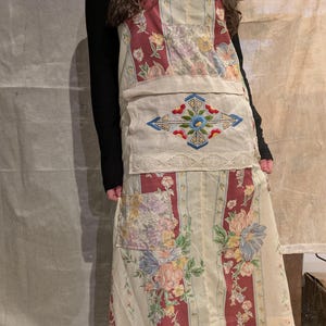 May include: A patchwork apron with a cream background, featuring floral and striped patterns in red, pink, and blue. The apron has a large pocket with an embroidered design and lace trim.