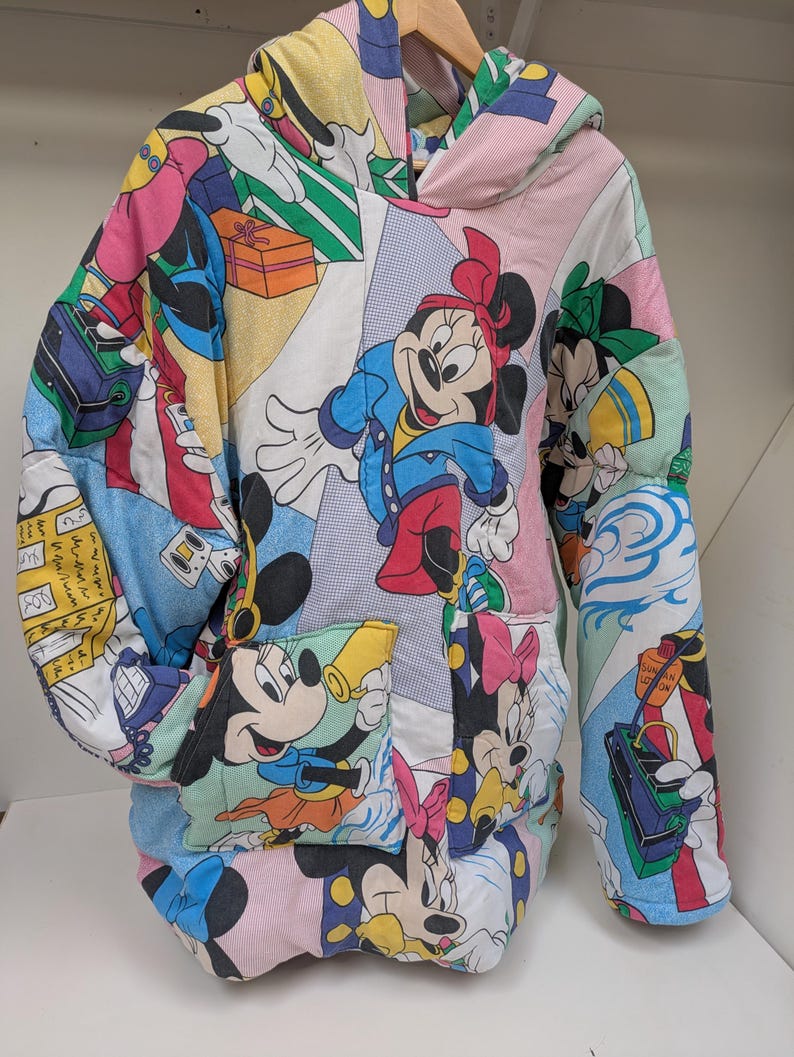 K&ouml;nnte beinhalten: Ein Patchwork-Hoodie mit Disney-Figuren wie Mickey und Minnie Mouse. Der Hoodie besteht aus bunten Stoffbahnen mit Cartoon-Grafiken, einer Kapuze und einer Vordertasche. Die &Auml;rmel sind lang und der Stil ist &uuml;bergro&szlig;.