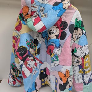 K&ouml;nnte beinhalten: Bunte Steppjacke mit Patchwork-Design, das verschiedene Bilder von Mickey und Minnie Mouse zeigt. Die Jacke hat eine Kapuze und h&auml;ngt an einem Holzb&uuml;gel. Die Farbpalette umfasst Rosa, Blau, Rot und Schwarz.