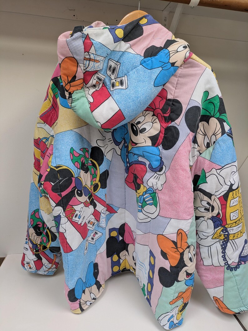 K&ouml;nnte beinhalten: Bunte Steppjacke mit Patchwork-Design, das verschiedene Bilder von Mickey und Minnie Mouse zeigt. Die Jacke hat eine Kapuze und h&auml;ngt an einem Holzb&uuml;gel. Die Farbpalette umfasst Rosa, Blau, Rot und Schwarz.