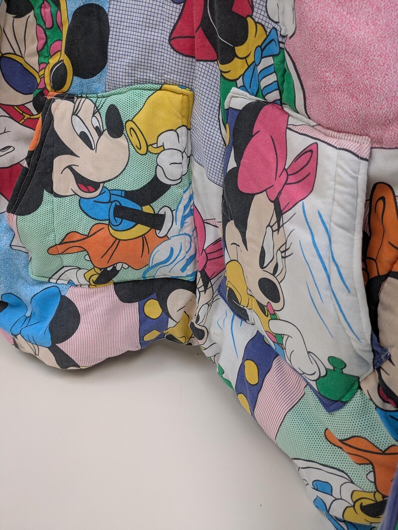 K&ouml;nnte beinhalten: Nahaufnahme eines Patchwork-Stoffs mit Micky und Minnie Maus. Der Stoff ist eine Mischung aus farbenfrohen Mustern und Drucken mit Bildern der ikonischen Zeichentrickfiguren. Die Taschen sind aus dem gleichen Stoff gefertigt.