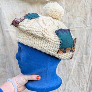 May include: Cream-colored knit hat with a cable knit design and a fluffy pom-pom on top. The hat features patches of teal and patterned fabric. The hat is displayed on a blue mannequin head.