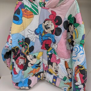 K&ouml;nnte beinhalten: Ein farbenfroher, gesteppter Kapuzenpullover mit einem Patchwork-Design, das verschiedene Disney-Zeichentrickfiguren, darunter Micky und Minnie Maus, zeigt. Der Kapuzenpullover hat eine Kapuze, Taschen und einen Rei&szlig;verschluss. Der Stoff ist eine Mischung aus Pastellfarben und Cartoon-Bildern.