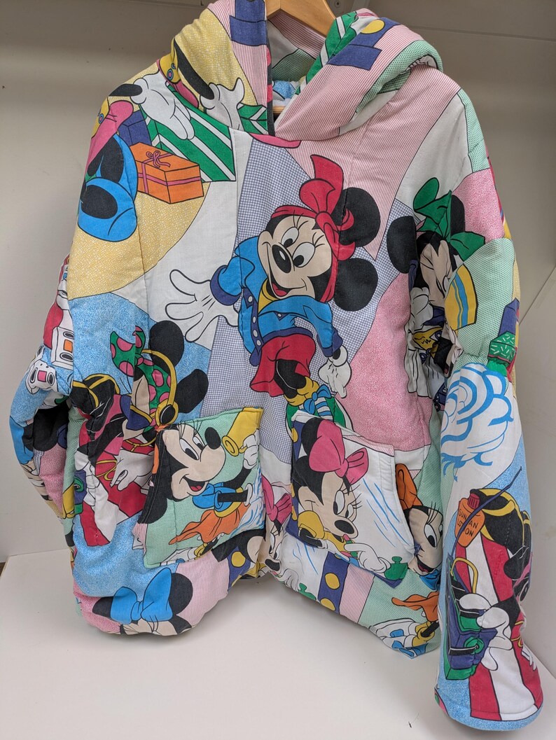 K&ouml;nnte beinhalten: Ein farbenfroher, gesteppter Kapuzenpullover mit einem Patchwork-Design, das verschiedene Disney-Zeichentrickfiguren, darunter Micky und Minnie Maus, zeigt. Der Kapuzenpullover hat eine Kapuze, Taschen und einen Rei&szlig;verschluss. Der Stoff ist eine Mischung aus Pastellfarben und Cartoon-Bildern.