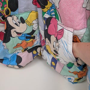K&ouml;nnte beinhalten: Eine Patchwork-Jacke mit Mickey- und Minnie-Mouse-Figuren. Die Jacke hat mehrere Taschen und ist aus einer Vielzahl von bunten Stoffen gefertigt. Das Design umfasst Bilder der Charaktere in verschiedenen Posen.