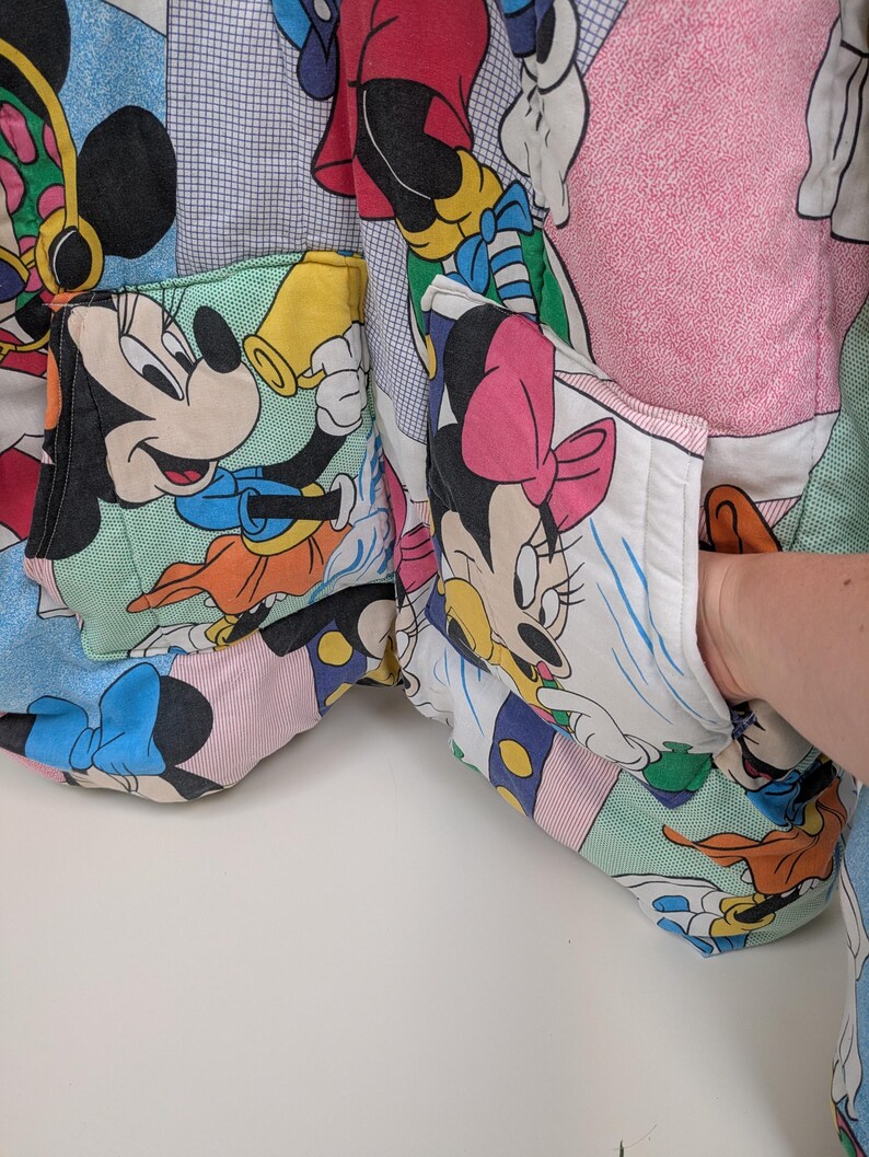 K&ouml;nnte beinhalten: Eine Patchwork-Jacke mit Mickey- und Minnie-Mouse-Figuren. Die Jacke hat mehrere Taschen und ist aus einer Vielzahl von bunten Stoffen gefertigt. Das Design umfasst Bilder der Charaktere in verschiedenen Posen.