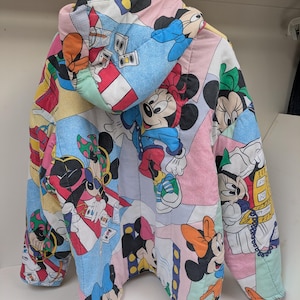K&ouml;nnte beinhalten: Eine farbenfrohe, gesteppte Jacke mit einem sich wiederholenden Muster von Mickey und Minnie Mouse. Die Jacke hat eine Kapuze und besteht aus verschiedenen pastellfarbenen Paneelen. Das Design enth&auml;lt Cartoon-Bilder und ist wahrscheinlich Vintage oder Retro.
