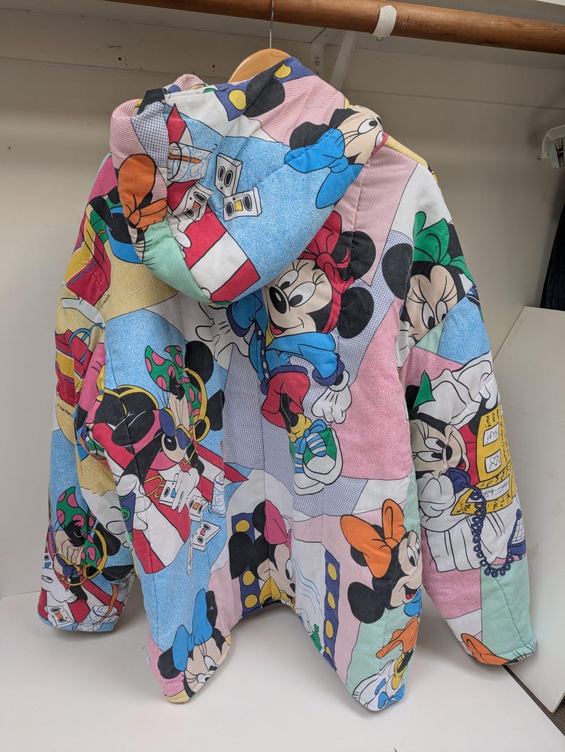 K&ouml;nnte beinhalten: Eine farbenfrohe, gesteppte Jacke mit einem sich wiederholenden Muster von Mickey und Minnie Mouse. Die Jacke hat eine Kapuze und besteht aus verschiedenen pastellfarbenen Paneelen. Das Design enth&auml;lt Cartoon-Bilder und ist wahrscheinlich Vintage oder Retro.