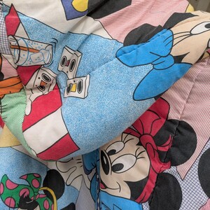 K&ouml;nnte beinhalten: Nahaufnahme einer bunten, gemusterten Bettdecke mit Zeichentrickfiguren. Das Design umfasst Bilder von Mickey und Minnie Mouse sowie andere Grafiken zum Thema Zeichentrick. Die Bettdecke hat ein blaues, rosa und wei&szlig;es Farbschema.