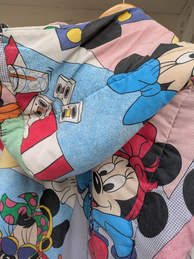 K&ouml;nnte beinhalten: Nahaufnahme einer bunten, gemusterten Bettdecke mit Zeichentrickfiguren. Das Design umfasst Bilder von Mickey und Minnie Mouse sowie andere Grafiken zum Thema Zeichentrick. Die Bettdecke hat ein blaues, rosa und wei&szlig;es Farbschema.