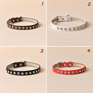 May include: Four pet collars in brown, white, black, and red, each featuring a row of clear circular rhinestones, a silver-toned metal buckle, and a light-colored interior lining. The collars are numbered one through four in the corners.