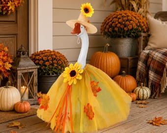 May include: A white goose figurine wearing a tan straw hat topped with a yellow sunflower, and a yellow tulle skirt decorated with orange and red fabric autumn leaves and a large yellow sunflower at the waist, featuring a bright orange beak.