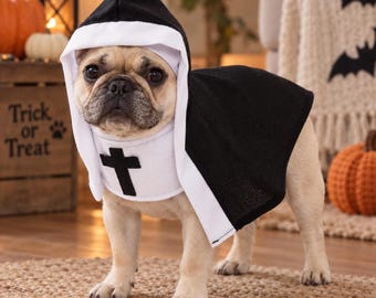 May include: A fawn-colored French Bulldog wearing a black and white nun costume. The outfit includes a black hooded cape with a white wimple and a black cross centered on the chest piece. The costume covers the head and back of the dog.
