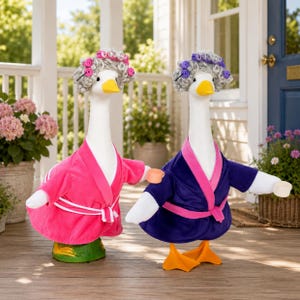 May include: Two decorative goose figurines, one in a pink robe and the other in a blue robe, both with curlers in their hair. The geese have white bodies, yellow beaks, and orange feet. They are holding small cups.