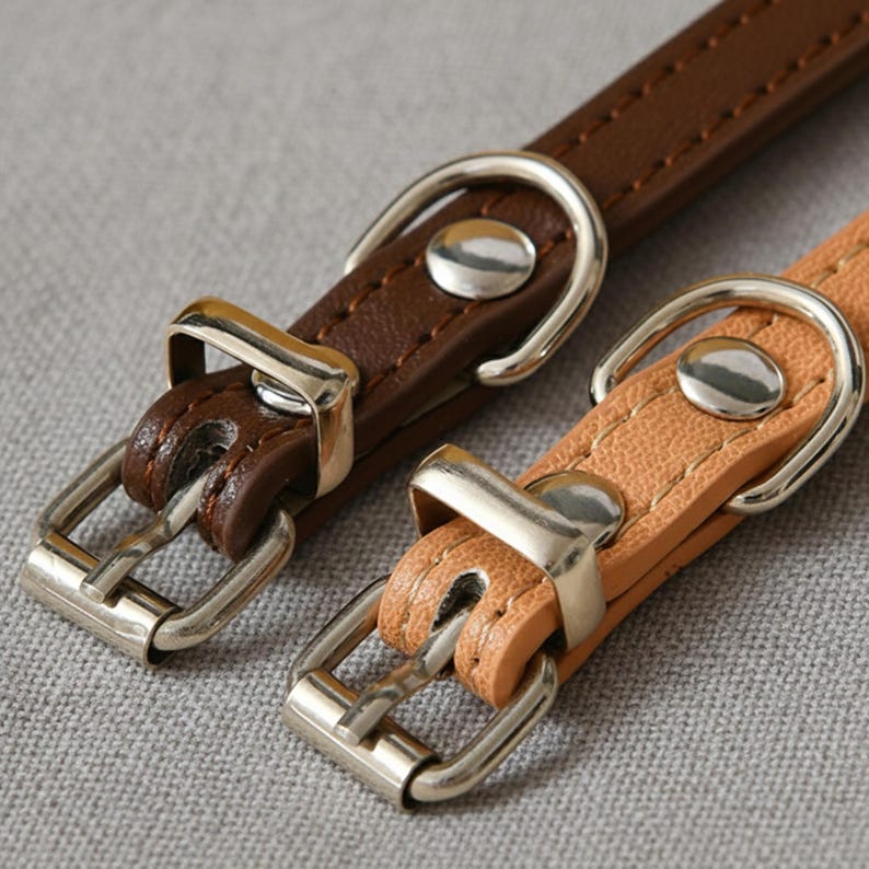 May include: Two narrow pet collars, one in dark brown and one in light tan, featuring textured material with visible stitching. Each collar includes a silver-toned metal buckle, a matching D-ring for leash attachment, and a fixed metal loop.