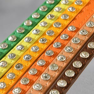 May include: A collection of five narrow, flat straps in green, yellow, light orange, dark orange, and brown. Each strap features a single row of evenly spaced, circular, clear faceted crystals set in silver-toned metal, with visible stitching.