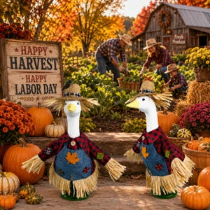 May include: Two goose figurines wearing straw hats, red and black plaid shirts, and blue denim overalls with leaf patches. Each figure has straw-like fringe at the base and sleeves, standing amidst pumpkins and autumn harvest decorations.