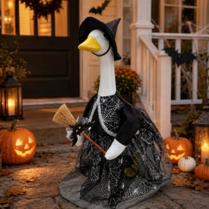May include: A white goose figurine dressed as a witch, wearing a black pointed hat and a black dress with a white spiderweb pattern and lace trim. The goose holds a small straw broom with a black bat decoration attached to the handle.
