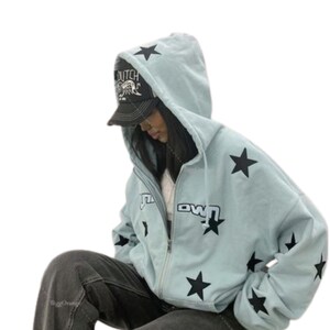 Puede incluir: Sudadera con capucha azul claro con estrellas negras y la palabra "Downtown" impresa. La sudadera con capucha se usa con una gorra de b&eacute;isbol negra. La sudadera tiene capucha.