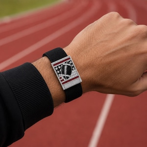 May include: A black watch band with a rectangular silver buckle featuring a football design. The buckle has a football in the center, surrounded by stars and red stripes. The watch is on a person's wrist.