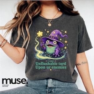 May include: A charcoal gray t-shirt featuring a graphic of a purple frog wearing a wizard hat and holding a glowing yellow star wand. Below the frog, the text reads: I cast Unflushable turd Upon ur enemies, surrounded by small yellow stars.