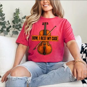 Könnte beinhalten: Ein kurzärmeliges T-Shirt in Rot meliert mit einer Grafik einer braunen Geige und einem Bogen. Über der Mitte der Geige befindet sich ein rechteckiges orangefarbenes Schild mit dem schwarzen Text "BOW, I REST MY CASE". Das Shirt hat einen Rundhalsausschnitt.