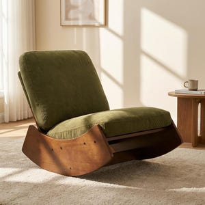 Pu&ograve; includere: Una moderna sedia a dondolo con struttura in legno e cuscini in velluto a coste verde oliva. La sedia ha una base curva per dondolare ed &egrave; posizionata su un tappeto chiaro. Un tavolino con una tazza &egrave; visibile sullo sfondo.
