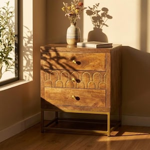 May include: A wooden nightstand with three drawers and brass hardware. The top two drawers have a carved arch design. The nightstand sits on a gold-colored metal frame. A vase of dried flowers and a book are on top.