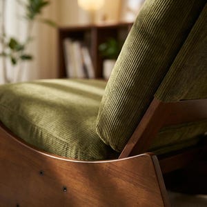Pu&ograve; includere: Primo piano di una sedia moderna di met&agrave; secolo con struttura in legno scuro e cuscini in velluto a coste verde oliva. La sedia ha schienale e seduta curvi, con venature del legno visibili. Il velluto a coste ha una trama a coste.