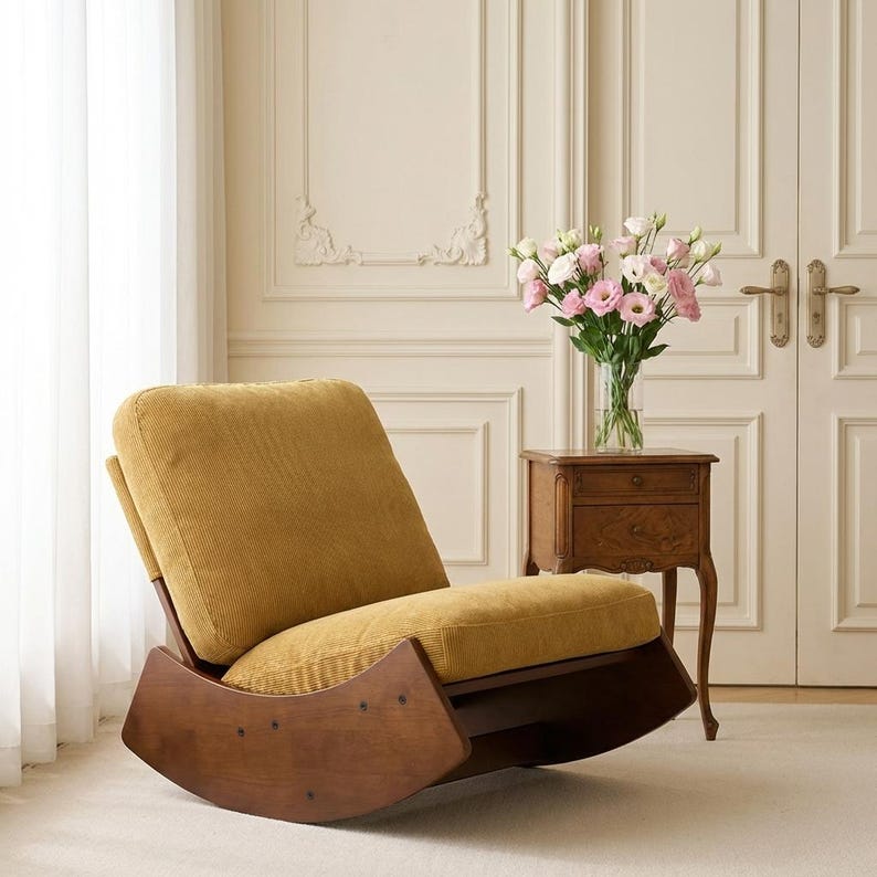 Pu&ograve; includere: Una poltrona a dondolo in velluto a coste giallo senape con struttura in legno. La sedia ha uno schienale alto e una seduta confortevole. I bilancieri in legno sono curvi. Un piccolo comodino in legno e un vaso di fiori rosa sono sullo sfondo.