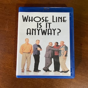 May include: A blue Blu-ray case with the title "Whose Line Is It Anyway?" prominently displayed. The cover shows six men in casual attire. The case is resting on a wooden surface.