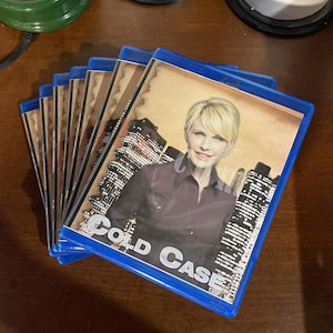 May include: A stack of Blu-ray discs with blue cases. The cover art features a woman with blonde hair in front of a cityscape, with the title "Cold Case" in white letters.