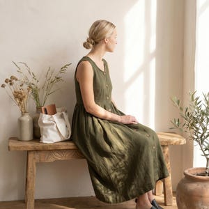 Puede incluir: Un vestido midi de color verde oliva sin mangas con cuello redondo y cierre frontal de botones. El vestido presenta un corpiño ajustado y una falda fruncida y fluida que cae por debajo de las rodillas, combinado con zapatos negros sin cordones.