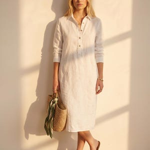 May include: A white long-sleeved midi shirt dress with a collar and a partial button-down front. The dress is paired with tan leather slide sandals and a woven straw tote bag featuring a green and white patterned silk scarf tied to the handle.
