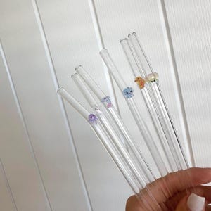 May include: A set of five clear glass drinking straws, each featuring a small, colorful, translucent animal-shaped charm attached to the side. The charms include a purple octopus, a white rabbit, a blue flower, an orange creature, and a yellow bear.