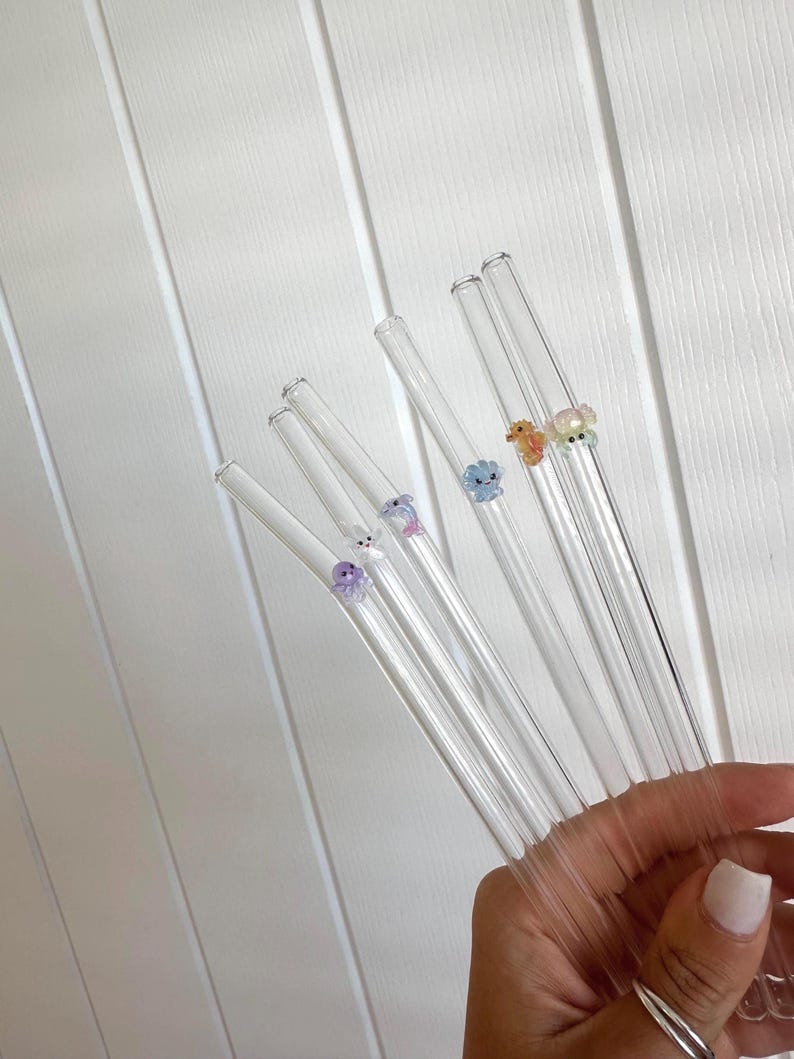 May include: A set of five clear glass drinking straws, each featuring a small, colorful, translucent animal-shaped charm attached to the side. The charms include a purple octopus, a white rabbit, a blue flower, an orange creature, and a yellow bear.