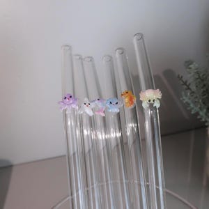 May include: A set of six clear glass drinking straws, each featuring a small, colorful, three-dimensional sea creature figurine attached to the side. The figures include a purple octopus, a white fish, a pink dolphin, a blue jellyfish, an orange seahorse, and a light pink crab.