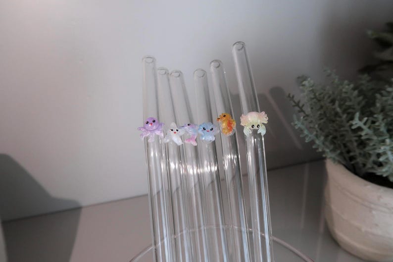 May include: A set of six clear glass drinking straws, each featuring a small, colorful, three-dimensional sea creature figurine attached to the side. The figures include a purple octopus, a white fish, a pink dolphin, a blue jellyfish, an orange seahorse, and a light pink crab.