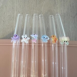 May include: Six clear glass drinking straws, each featuring a small, colorful, three-dimensional sea creature figurine attached to the side. The figures include a purple octopus, a white starfish, a pink dolphin, a blue shell, an orange seahorse, and a crab.